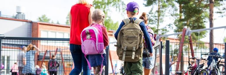 Lapset kävelee koulun pihaan reput selässä.