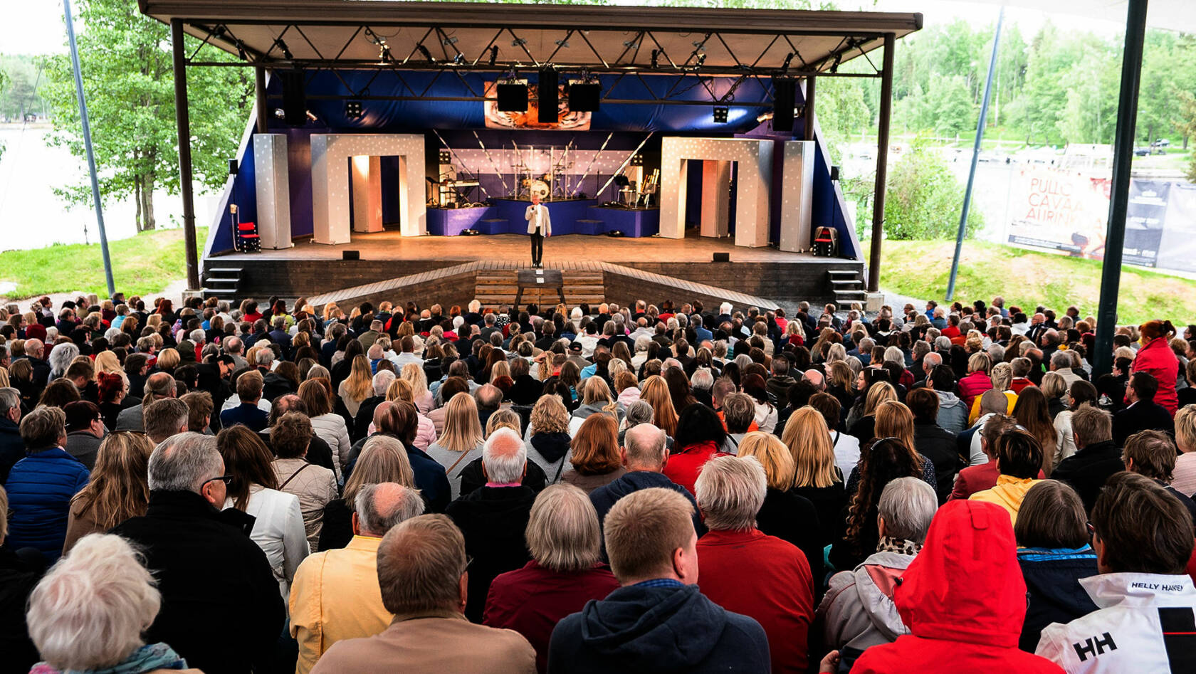 Apianlahden kesäteatterin katsomo, jossa on yleisöä ja lavalla seisoo Heikki Paavilainen.