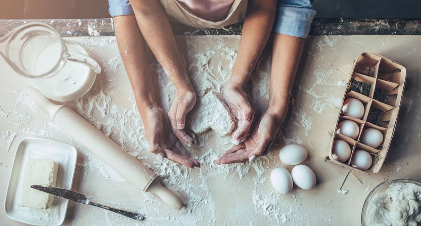Kahdet kädet leivovat sydämenmuotoista pullaa. Pöydällä myös voita, munia ja jauhoja.