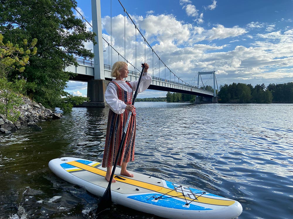 Nainen, jolla on yllään Sääksmäen kansallispuku sup-lautailee Sääksmäen sillan lähellä järvellä.