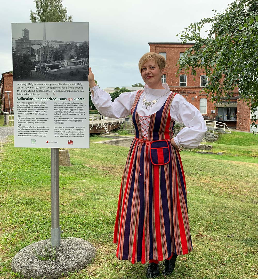 Nainen, jolla on yllään Sääksmäen kansallispuku, seisoo valokuvakyltin vieressä Myllysaaren museon pihassa.