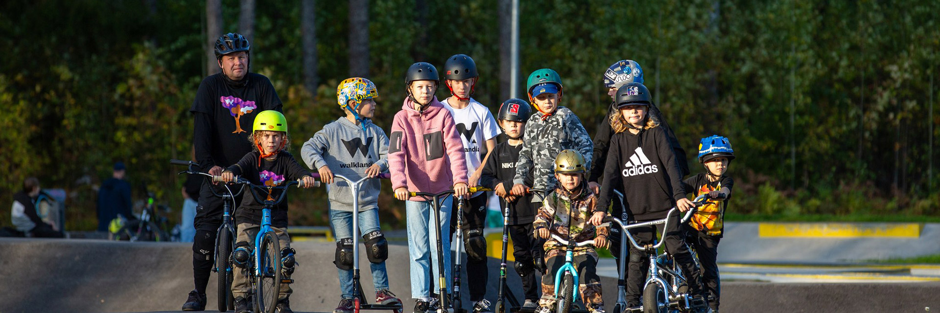Lapsia ja aikuinen pyörien sekä potkulautojen kanssa pumptrack-radalla Walklandiassa.