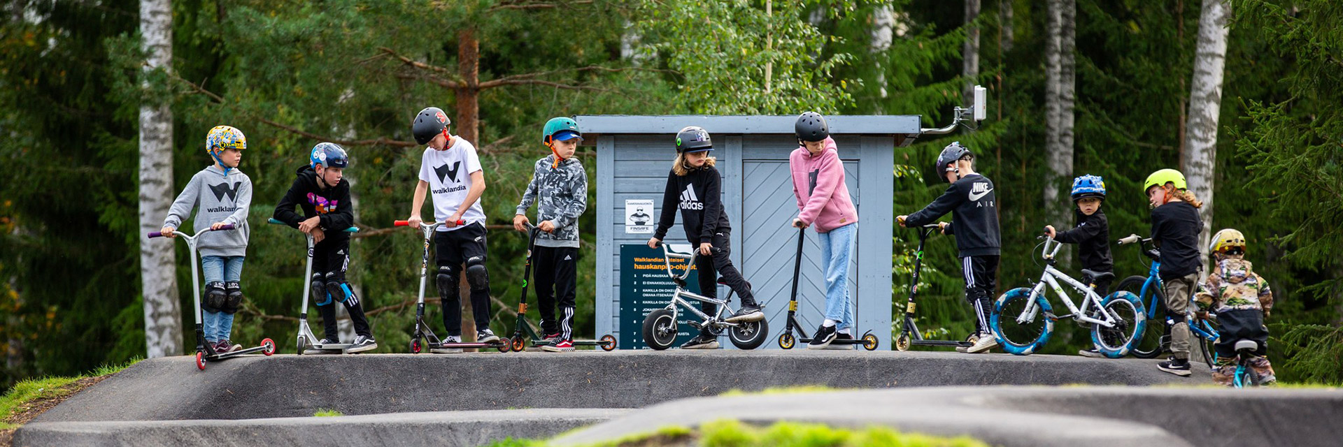 Kids on the pump track with their bikes and kick scooters.