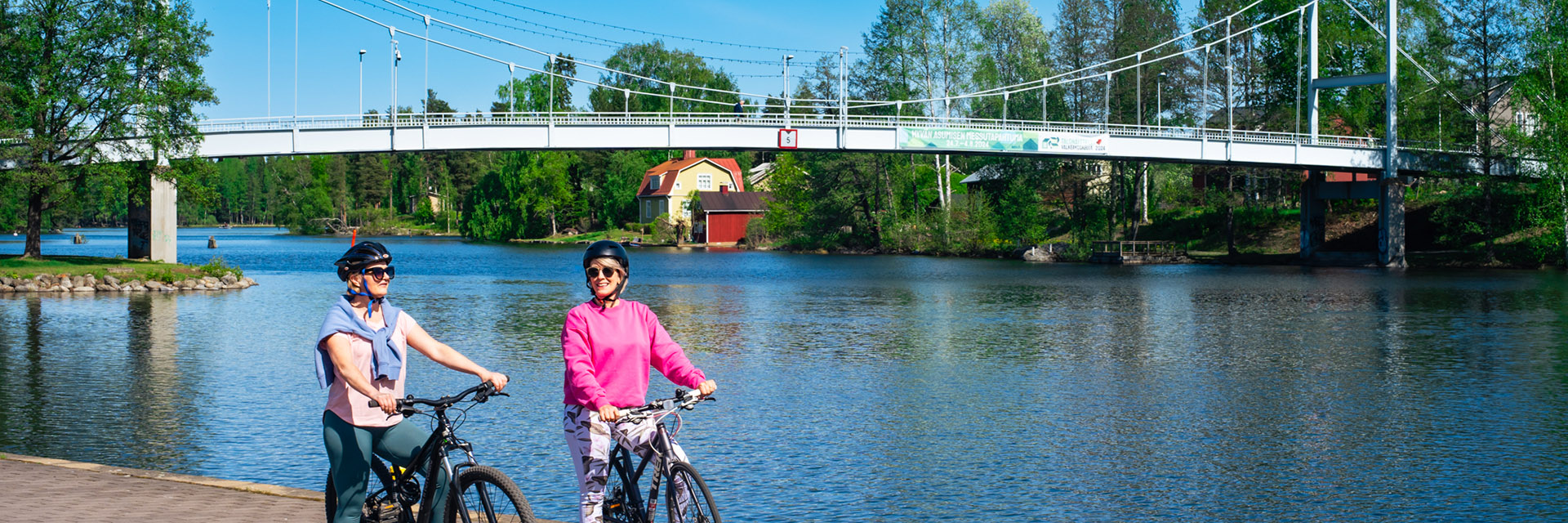 Kaksi naista pyöräilee Valkeakosken keskustassa järven rannassa.