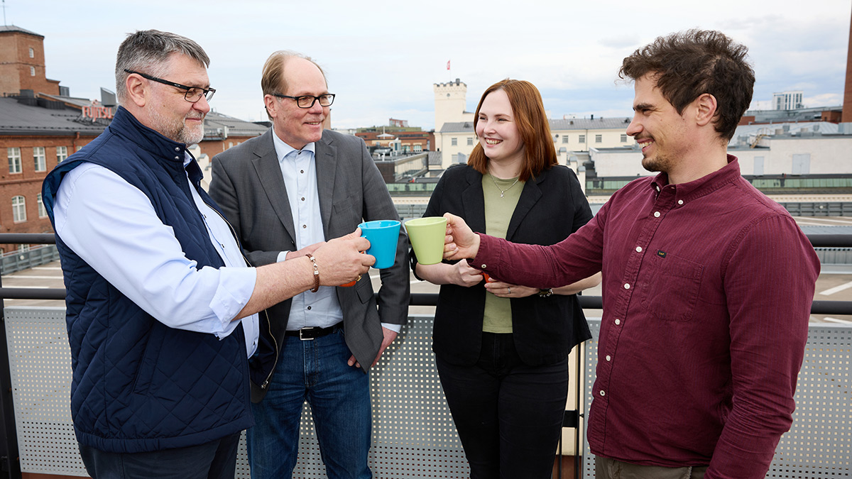 Neljä henkilöä seisovat ulkona parvekkeella ja kippistävät kahvimukeilla.