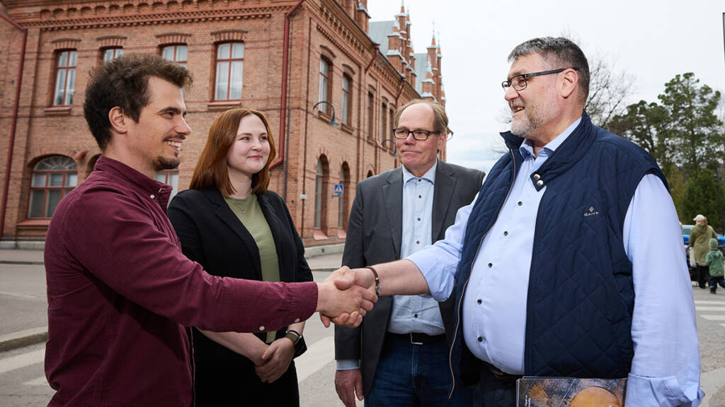 Neljä henkilöä seisovat vastakkain, joista kaksi kättelevät toisiaan ja kaksi muuta katsovat kättelijöitä.