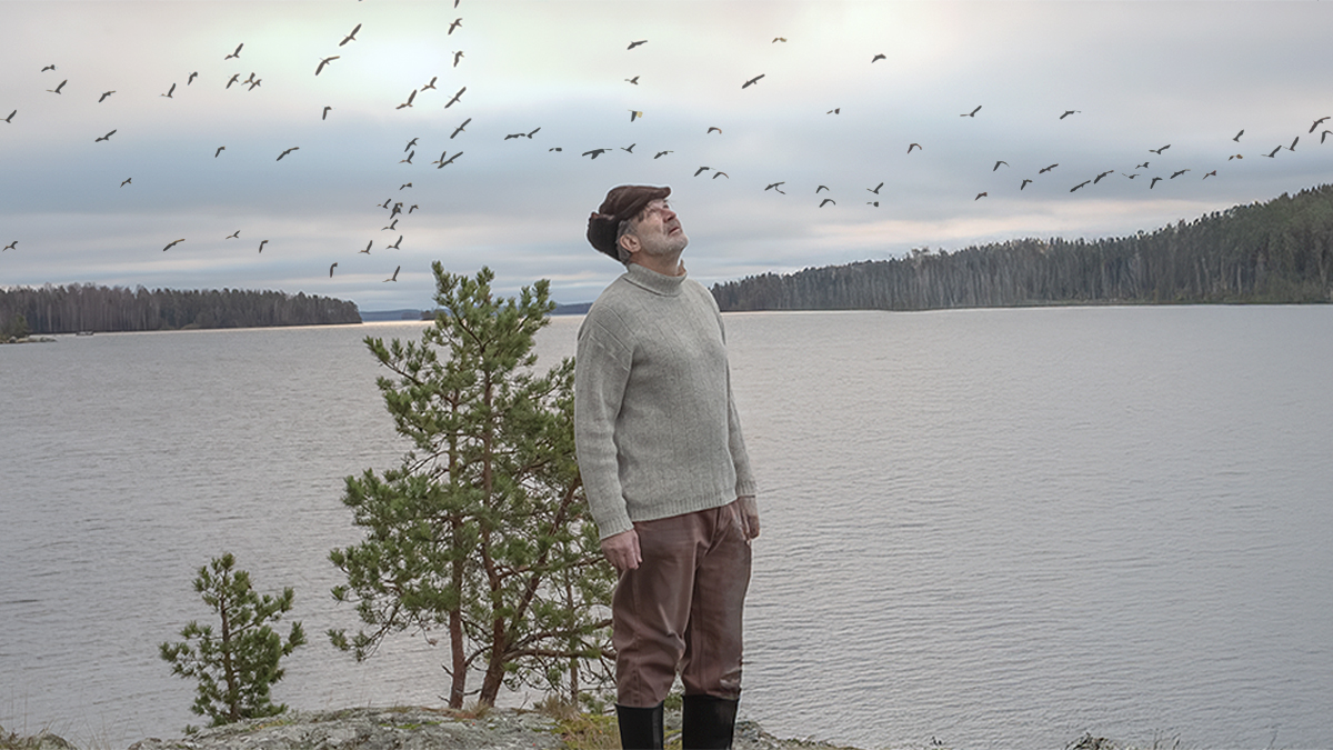 Mies seisoo kalliolla järven vieressä ja katselee taivaalla lentäviä lintuja.