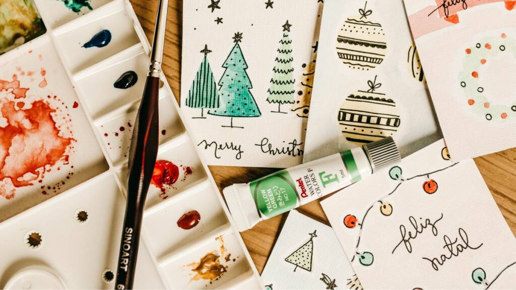 Joulukortteja, vesivärejä, sivellin ja liimaputkilo pöydällä.