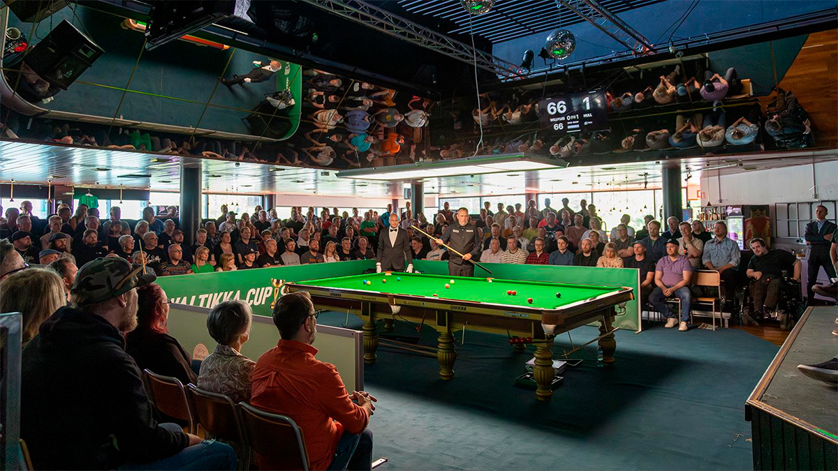 Biljardi-kilpailuissa pelaajia pöydän äärellä ja yleisöä katsomossa.