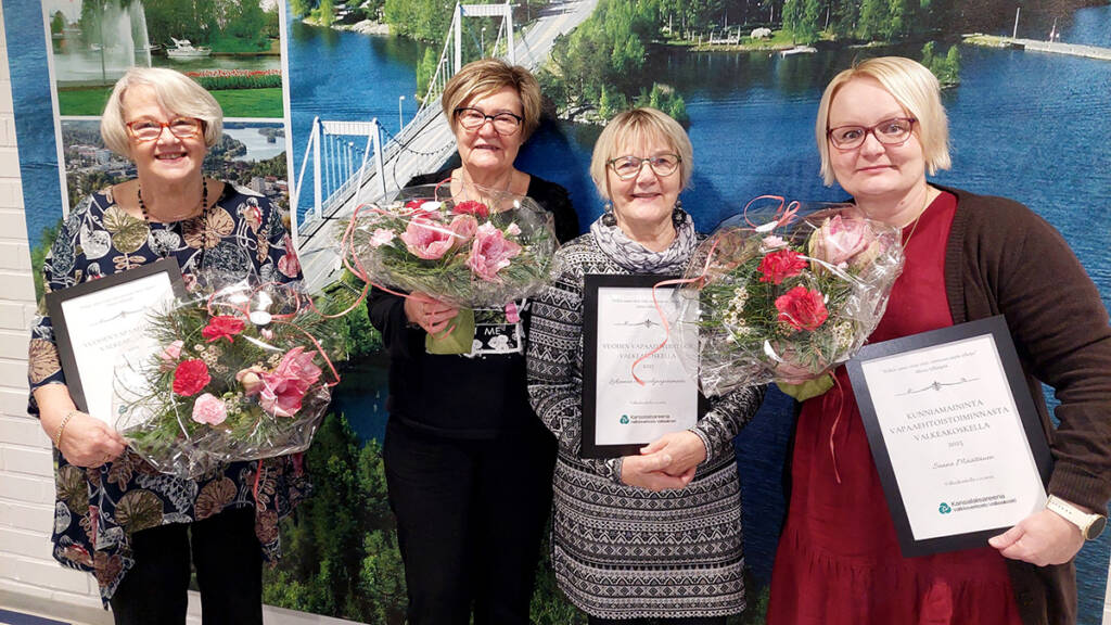 Neljä henkilö seisovat rinnakkain ja kolme heistä pitelevät kukkakimppuja ja yksi pitelee palkintodiplomia.