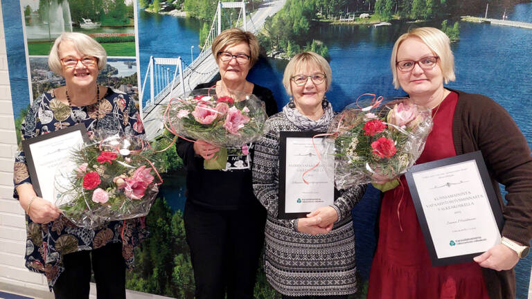 Neljä henkilö seisovat rinnakkain ja kolme heistä pitelevät kukkakimppuja ja yksi pitelee palkintodiplomia.