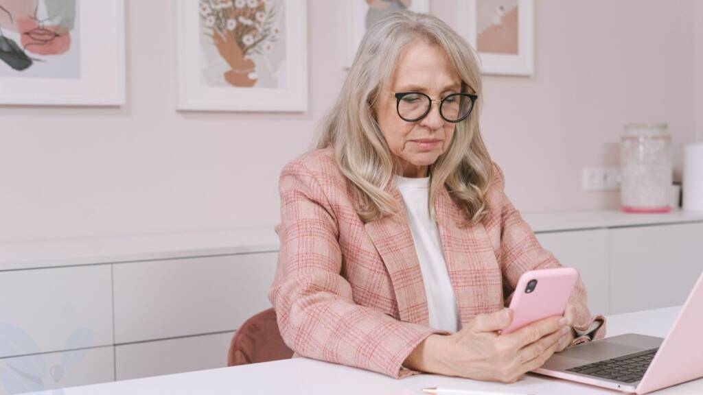 Nainen istuu pöydän ääressä katsellen puhelintaan ja tietokone on hänen edessään.