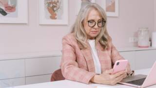 Nainen istuu pöydän ääressä katsellen puhelintaan ja tietokone on hänen edessään.