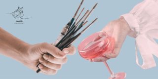 Vasemmalla käsi pitää siveltimiä ja oikealla kädessä on viinilasi.