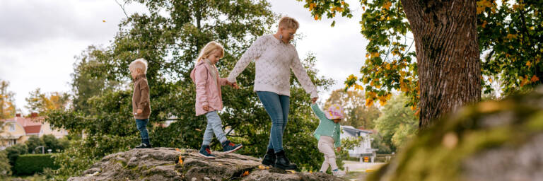 Äiti ja kolme lasta kiipeilevät käsikädessä kivien päällä puistossa.