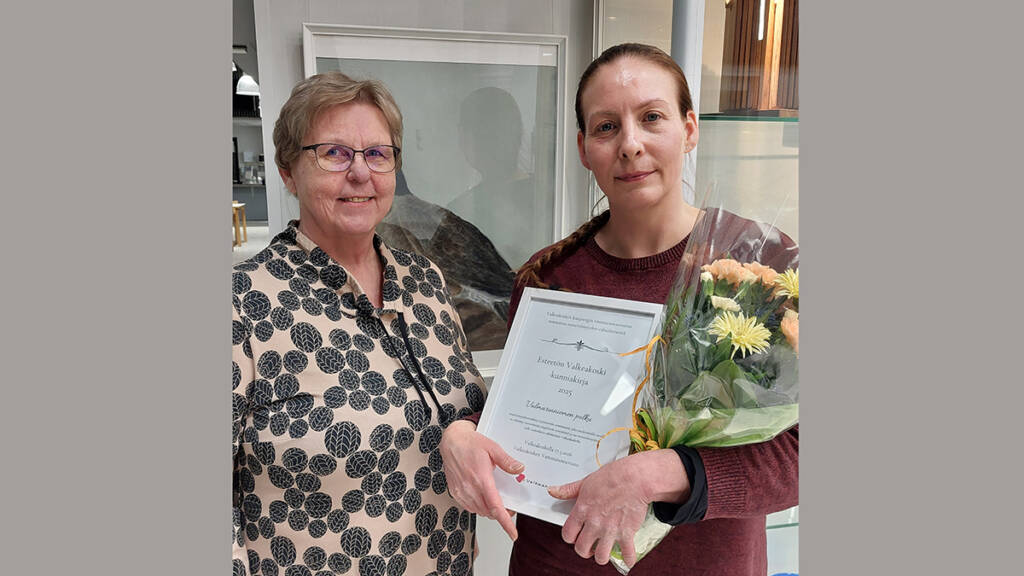 Kaksi henkilöä valokuvassa, toinen heistä pitelee käsissään kunniakirjaa ja kukkakimppua.