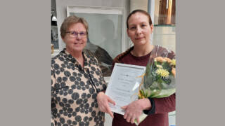 Kaksi henkilöä valokuvassa, toinen heistä pitelee käsissään kunniakirjaa ja kukkakimppua.