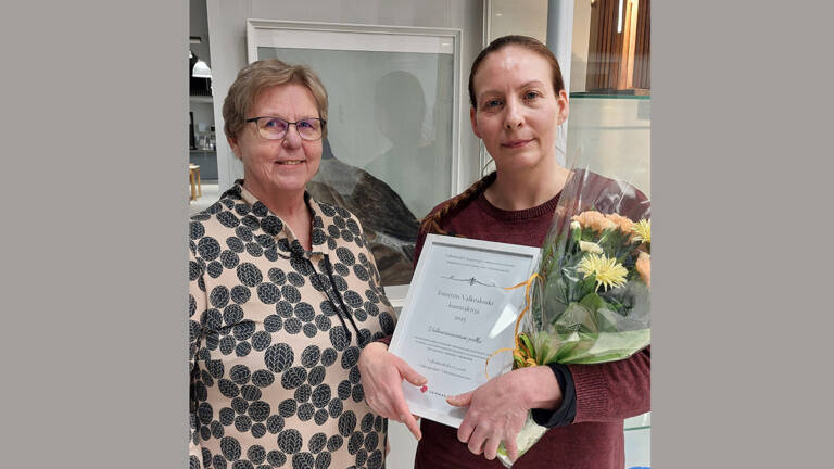 Kaksi henkilöä valokuvassa, toinen heistä pitelee käsissään kunniakirjaa ja kukkakimppua.