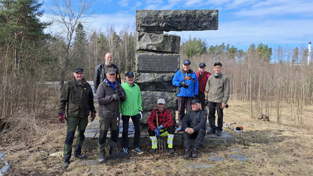 Yhdeksän miestä seisoo muistomerkin edustalla, taustalla puistomaisema.