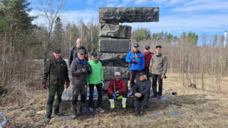 Yhdeksän miestä seisoo muistomerkin edustalla, taustalla puistomaisema.