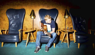 Mies istuu tuolissa ja soittaa kitaraa.