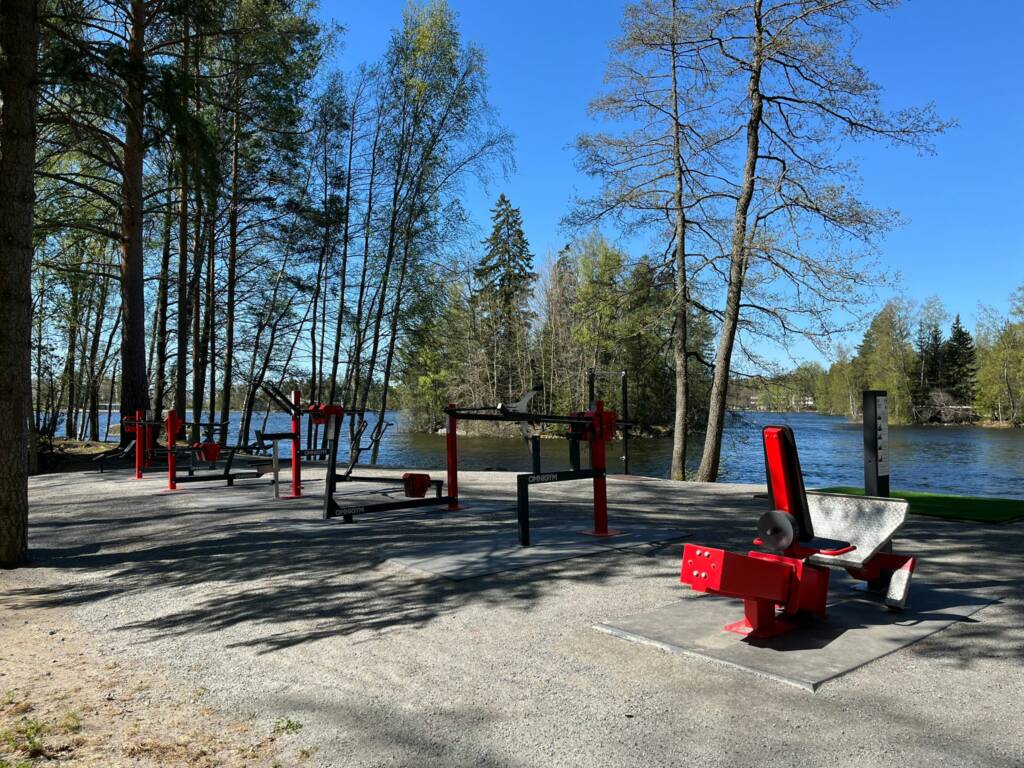 Ulkona järven rannalla erilaisia kuntosalilaitteita.