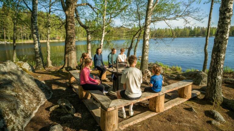 Perhe istuu nuotiopaikalla kesällä, taustalla näkyy järvi ja puita.
